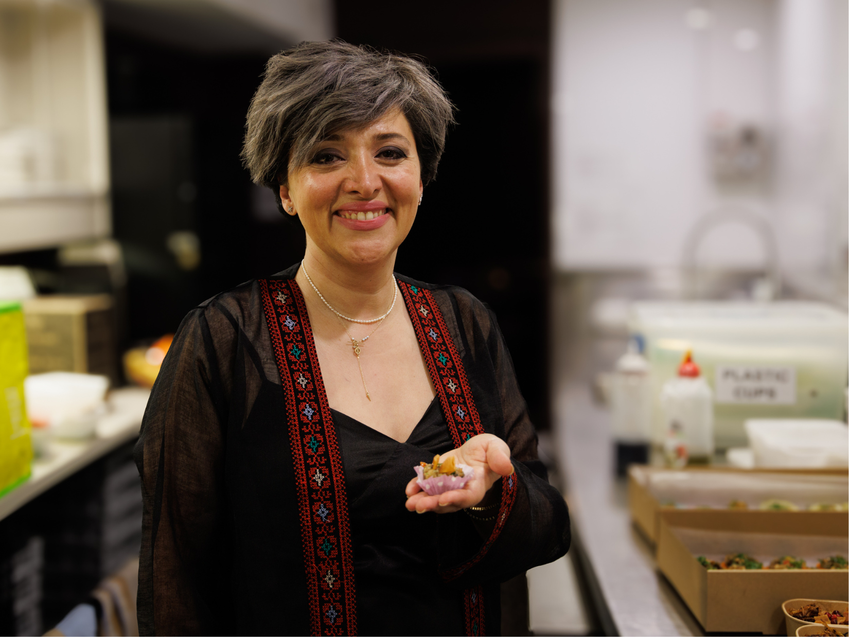 Image supplied: Racha, from Racha's Syrian Kitchen; A woman with short, gray hair smiles warmly while holding a small dish in her hand. She is wearing a black top with intricate red and blue embroidery. The background shows a kitchen setting with various items on the counter.